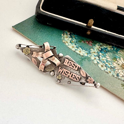 Victorian 1900 antique sterling silver and rose gold sweetheart brooch with the words 'BEST WISHES' in raised lettering, styled on a floral green card next to a vintage black case.
