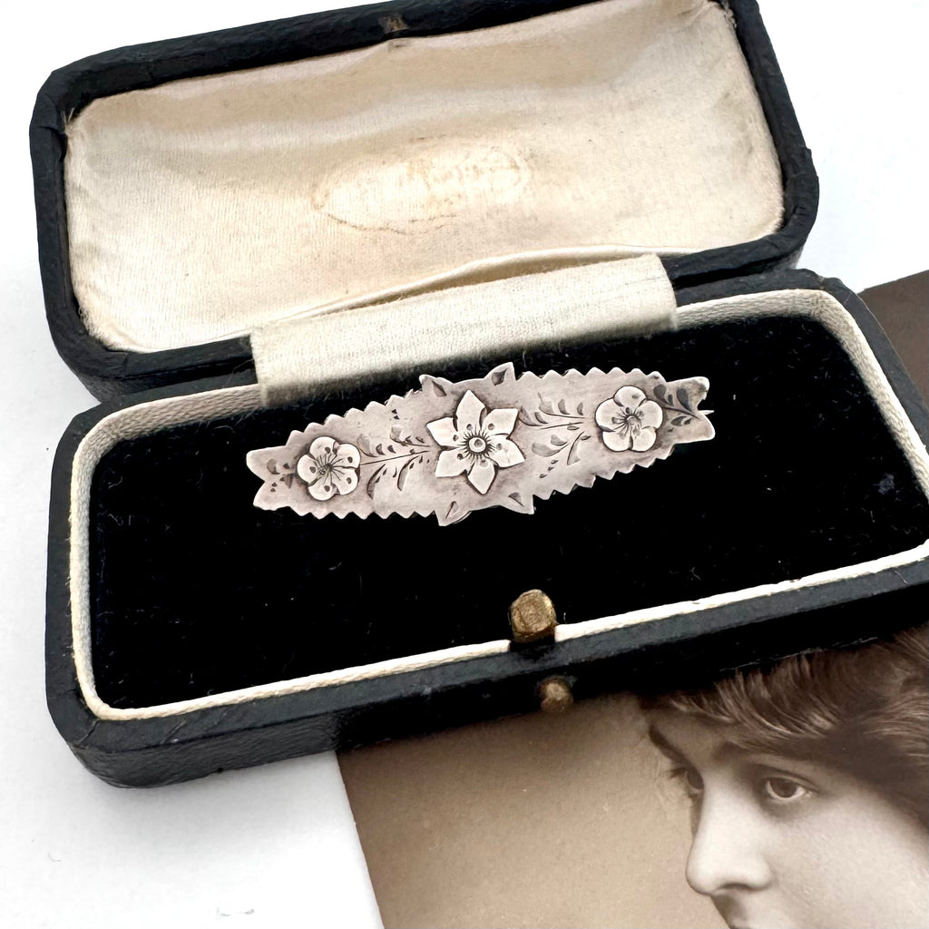 Victorian silver floral sweetheart brooch displayed inside an open black velvet jewelry box with a cream lining, with a vintage portrait card partially visible underneath.
