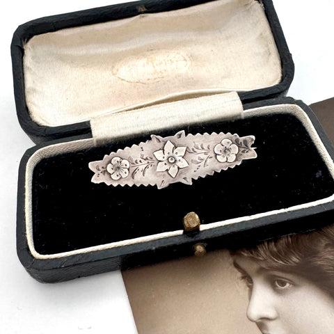 Victorian silver floral sweetheart brooch displayed inside an open black velvet jewelry box with a cream lining, with a vintage portrait card partially visible underneath.