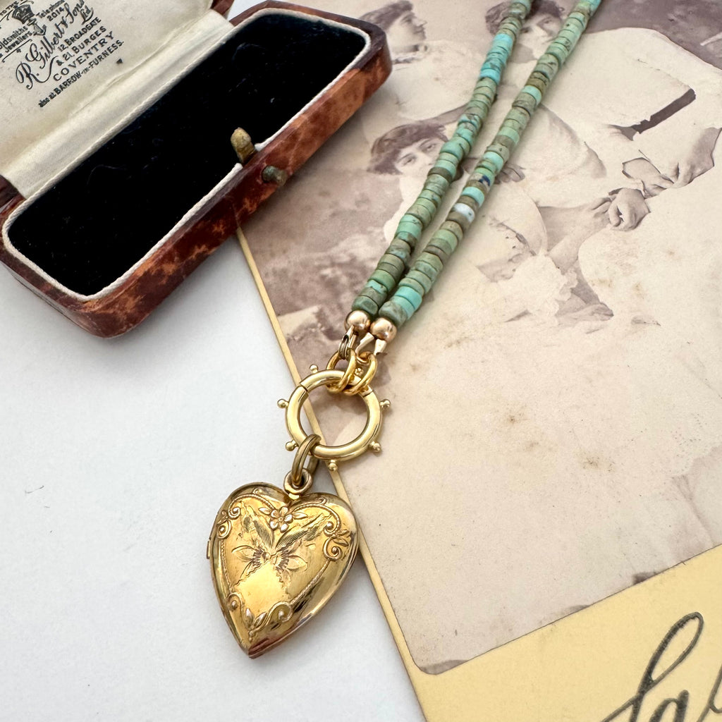 A vintage Carl Art gold heart-shaped locket pendant and a beaded turquoise-blue chain, displayed next to an open brown jewelry box on a vintage printed card.