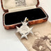 An antique sterling silver star-shaped watch fob with a circular center medallion engraved with 'SCHOOL OF ARMS' displayed on a vintage photograph next to an old wooden box.