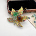 A vintage European hand wired gold-toned brooch shaped like a lotus flower with green, amber, and multicolor rhinestone accents, displayed near an old jewellery box and vintage postcard