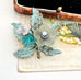A vintage 1930's European handwired floral brooch featuring a central frosted white flower, a translucent teal gemstone, a pale pink crystal, and glittery teal leaves, displayed on a textured vintage card.