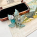 A vintage 1930's European handwired floral brooch with a translucent milky white flower, turquoise blue bead, and glittery mint green leaves, displayed next to an open vintage jewelry box on a textured beige surface.