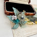 A decorative vintage 1930's handwired brooch featuring glittery turquoise-green leaves, a translucent white flower, a teal gemstone, and a small pale pink stone, displayed in front of an open jewelry box on a vintage-style card.