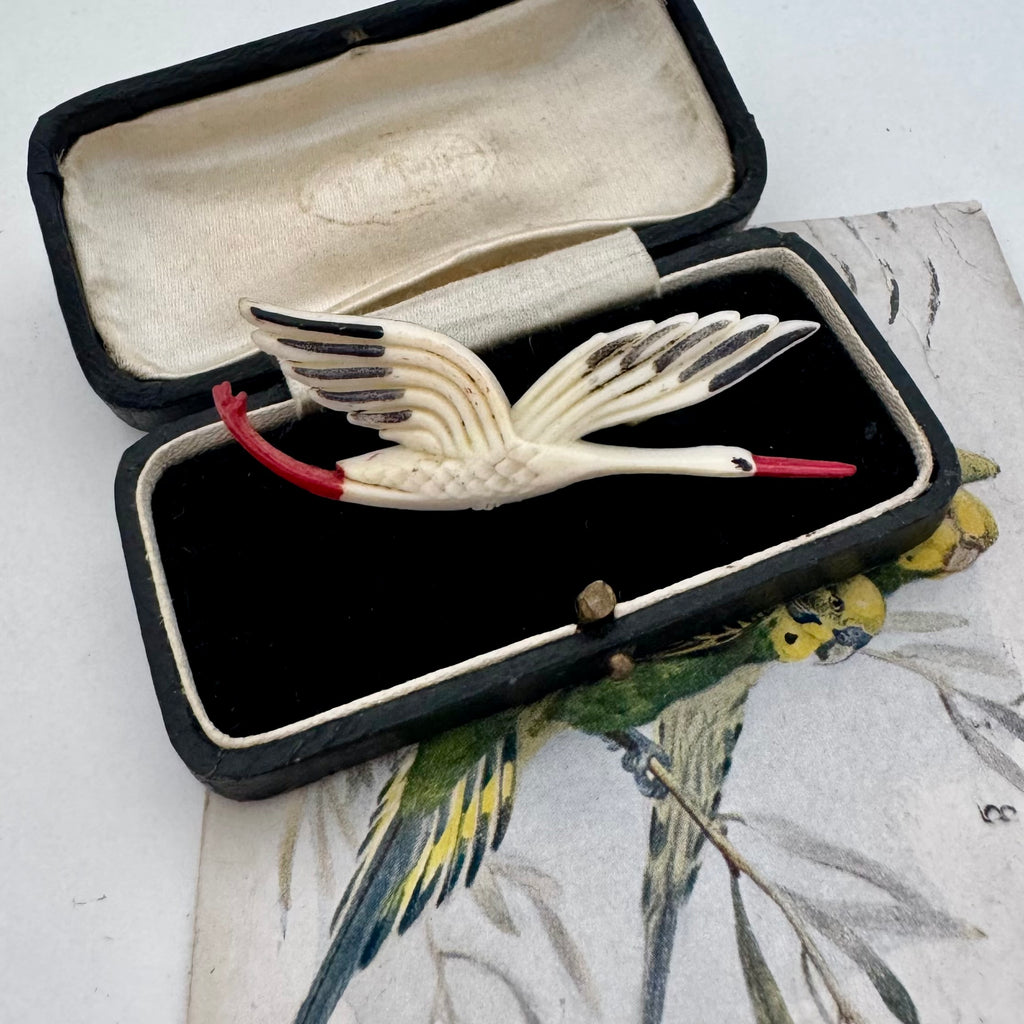 An antique stork brooch with cream-colored feathers, black wing tips, and a red beak and tail, displayed inside an open vintage black velvet jewelry box.