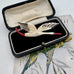 An antique stork brooch with cream-colored feathers, black wing tips, and a red beak and tail, displayed inside an open vintage black velvet jewelry box.