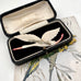 A vintage 1930's flying stork brooch or pin with cream-colored body and wings, black feather accents, and a red beak, displayed on a printed card with an open dark case in the background. 