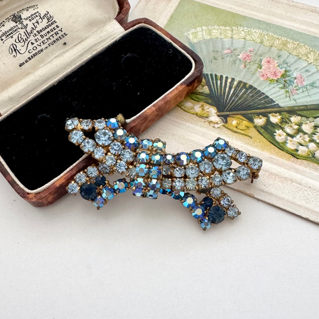 A vintage-style rhinestone brooch in a bow shape, featuring light blue, dark blue, and iridescent blue stones set in gold-toned metal, displayed next to its open vintage presentation box on a light surface.