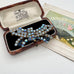 A vintage rhinestone brooch with blue and clear stones, displayed in an open wooden jewelry box next to a decorative fan illustration on a white background.