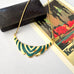 Gold necklace with a scalloped Art Deco style pendant featuring green inlay, displayed on a light gray surface next to a vintage landscape card and a dark textured box.