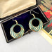 A pair of gold-tone hoop earrings with concentric circular enamel detailing in teal and cream, displayed inside an open black jewelry box with a dark purple velvet lining.