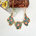 A vintage 1980's gold-tone statement necklace with three teardrop-shaped enamel pendants featuring a teal, pink, and gold floral pattern, displayed on a white surface with a vintage card and brown box as props.