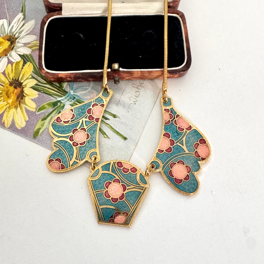 A vintage 1980's gold-tone necklace with three Art Nouveau style enamel pendants in teal, pink, and burgundy, displayed on a white surface next to a vintage flower illustration and a small jewelry box.