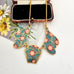 A vintage 1980's gold-tone necklace with three Art Nouveau style enamel pendants in teal, pink, and burgundy, displayed on a white surface next to a vintage flower illustration and a small jewelry box.