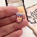 Small shield-shaped pendant with 'NORWICH' text held between fingers, with a decorative background.