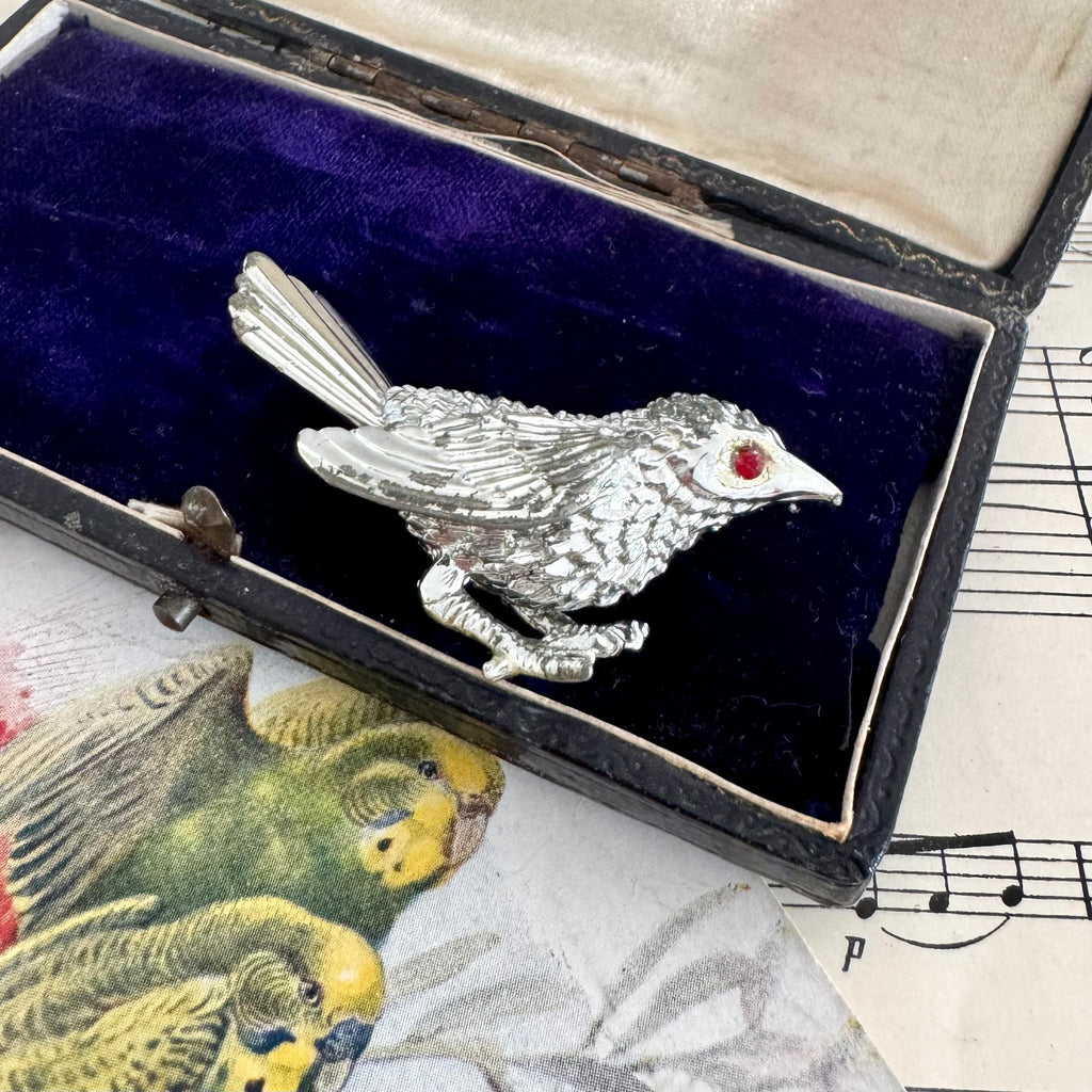 Vintage Sarah Coventry silver bird-shaped brooch with a red gemstone in an open jewelry box on sheet music.