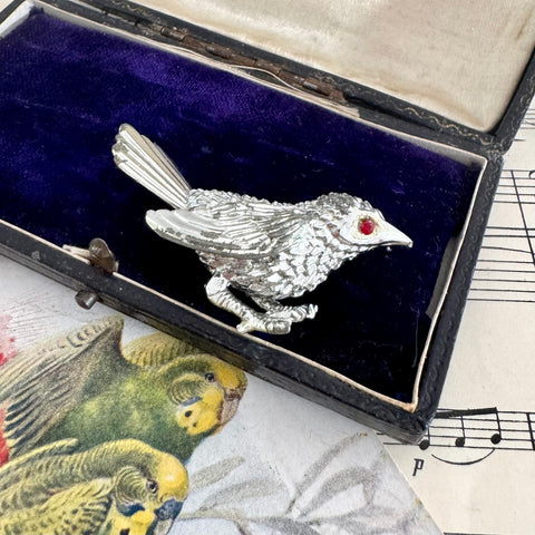 Vintage Sarah Coventry silver bird-shaped brooch with a red gemstone in an open jewelry box on sheet music.