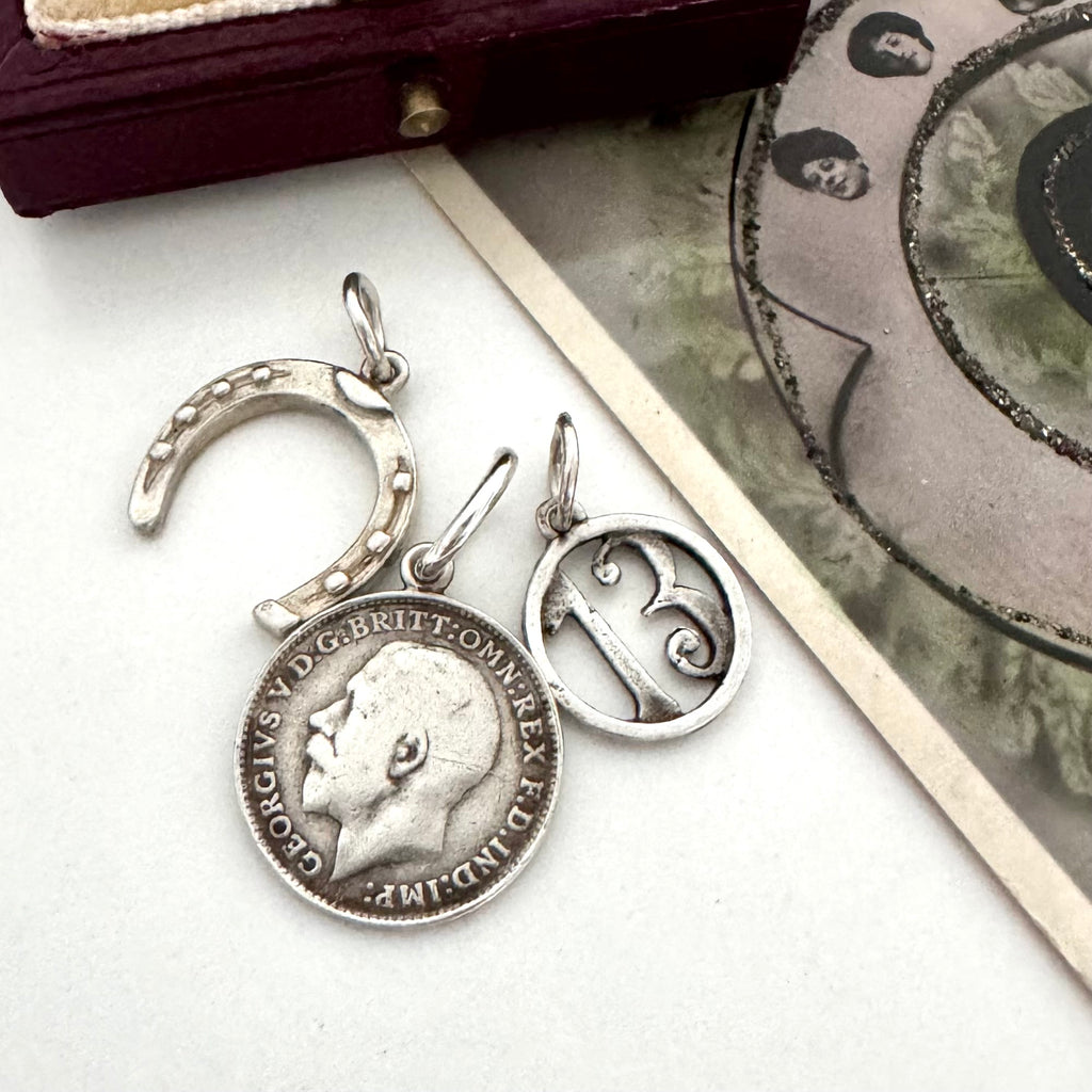 A vintage silver horseshoe charm, no 13 charm and a 1918 British threepence on a white background with an old postcard in the corner.