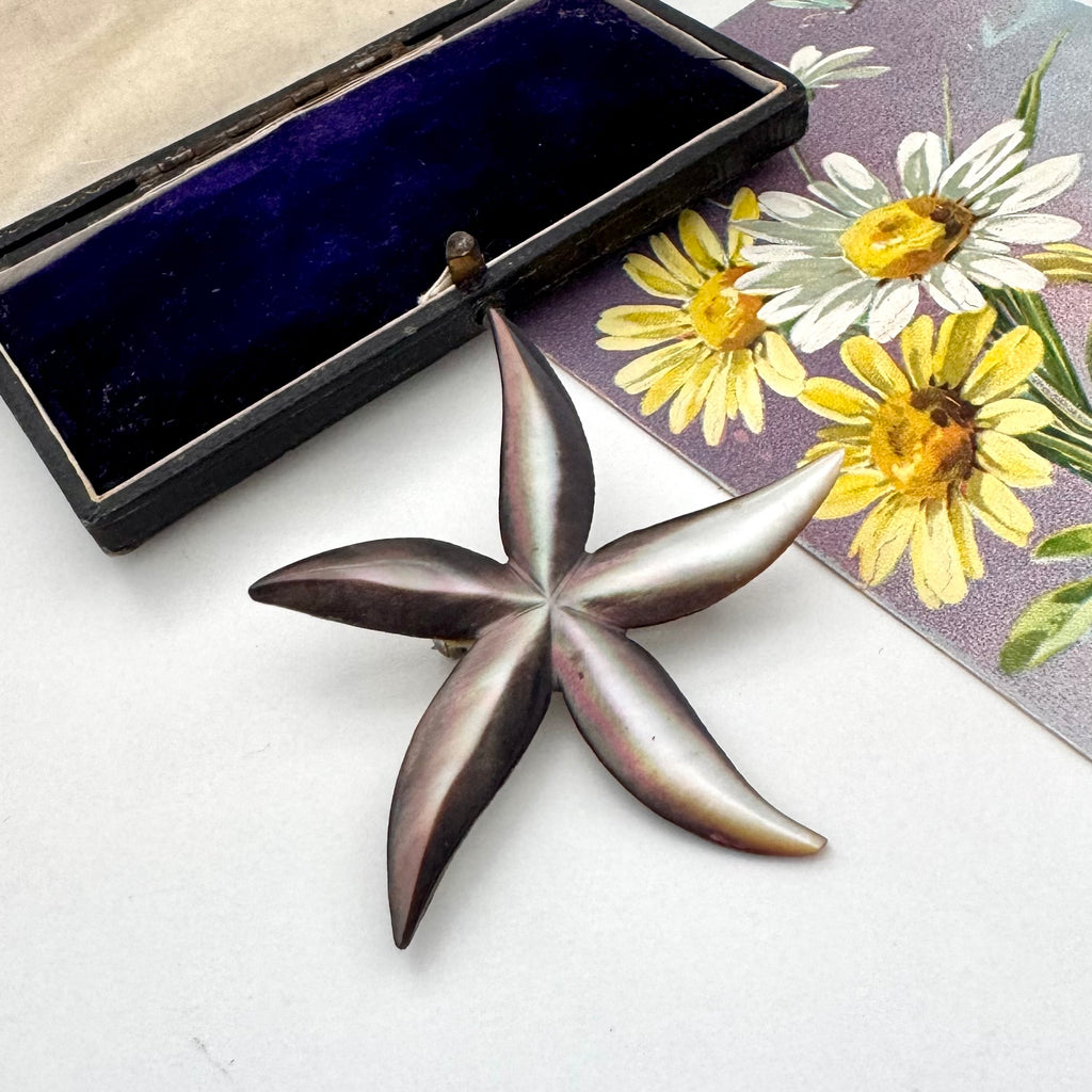Vintage starfish-shaped black pearl shell brooch on a white surface with a floral card in the background