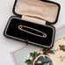 Gold collar safety pin in an open black jewelry box with a decorative card in the background