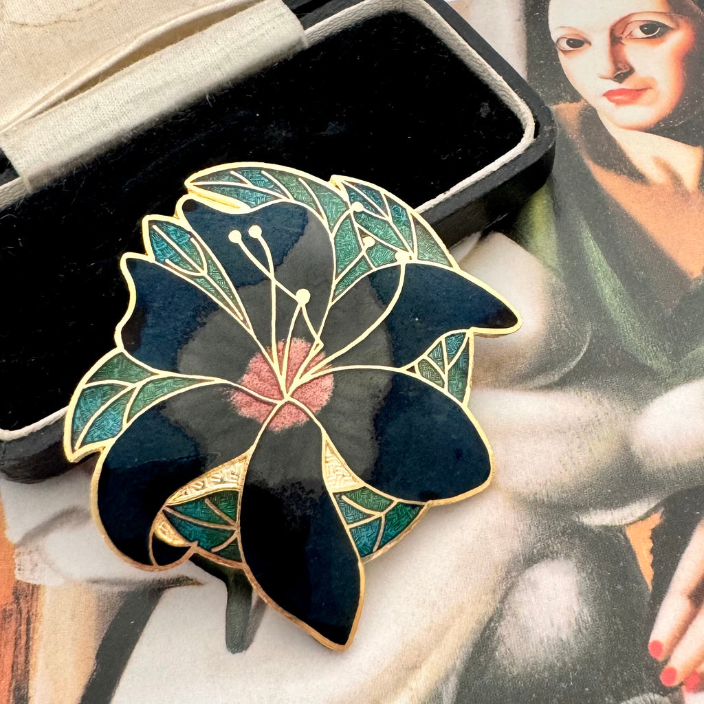A  vintage floral cloisonne enamel pin with black petals, green leaves, and gold outlines, displayed next to a black presentation box on a vintage-style painting background.