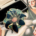 A  vintage floral cloisonne enamel pin with black petals, green leaves, and gold outlines, displayed next to a black presentation box on a vintage-style painting background.
