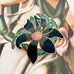 A  vintage floral cloisonne enamel pin with black petals, green leaves, and gold outlines, displayed on a vintage-style painting background.
