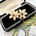 Vintage double daffodil flower brooch displayed inside an open vintage black velvet jewelry box with a beige lining, set against a background featuring a parrot illustration.