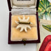 White flower-shaped brooch in a wooden box with 'Melbourne' branding, surrounded by botanical illustrations.