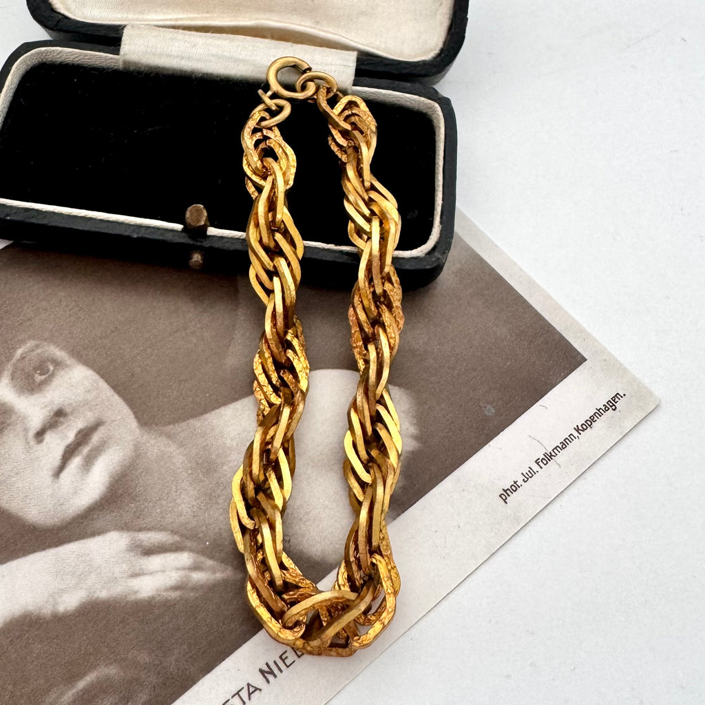 A chunky gold chain bracelet with a thick, twisted rope-like link design, displayed inside an open black jewelry box on top of a vintage black-and-white photograph.