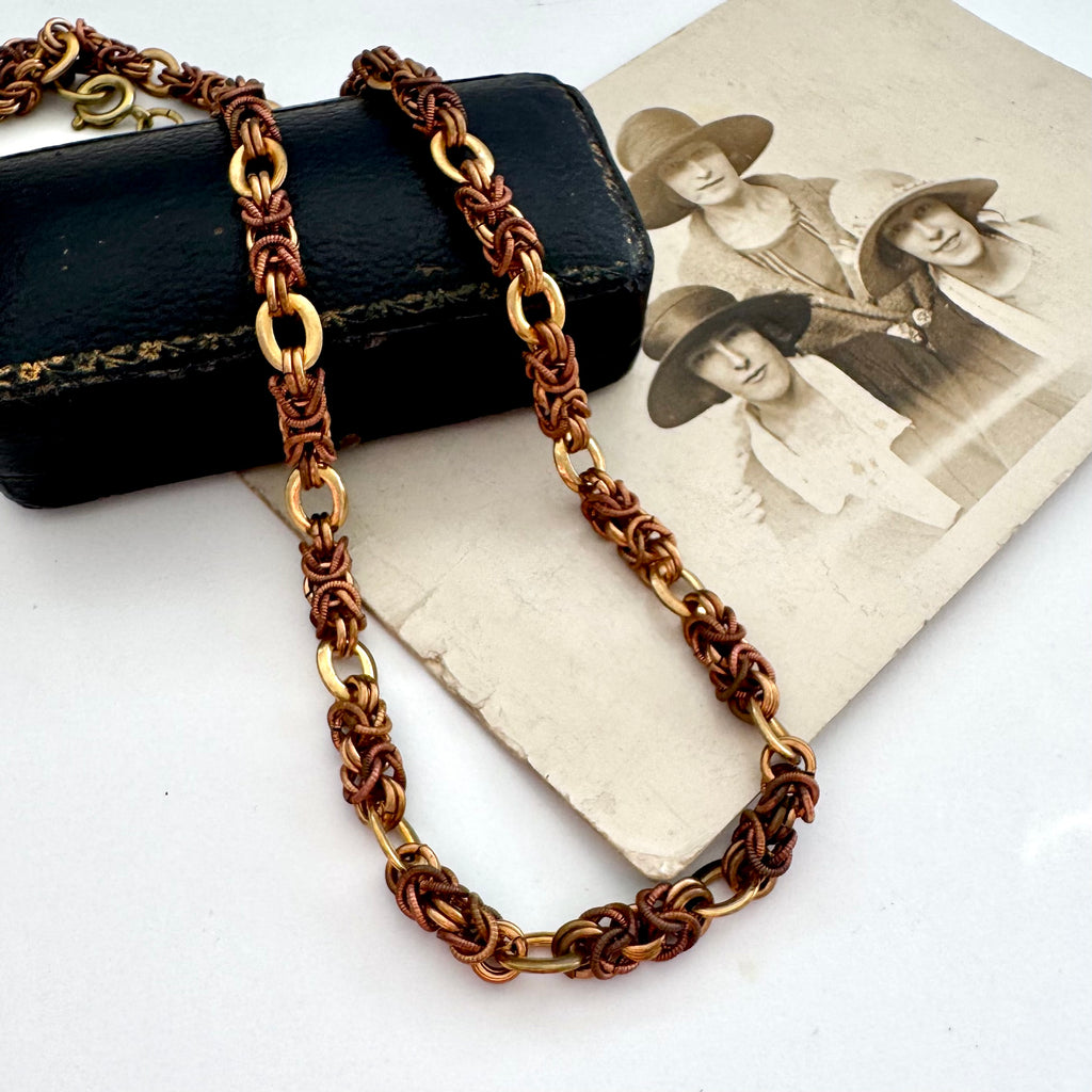 A vintage-Jakob Bengel Byzantine chain necklace with interwoven copper and gold links, draped over a black leather case and an old sepia-toned family photograph on a white surface.