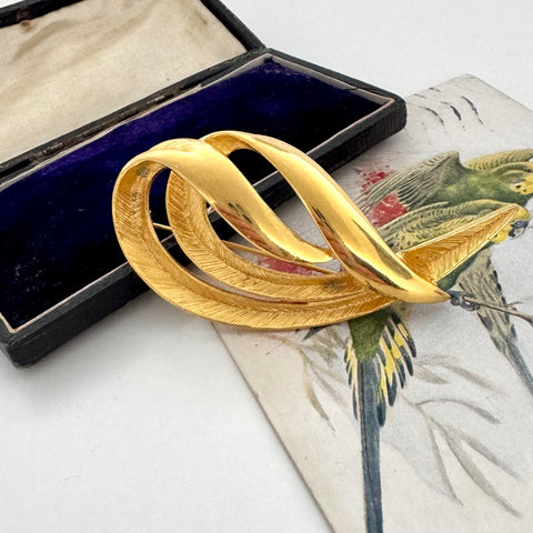 Vintage gold feather brooch on a decorative surface with an open jewelry box in the background