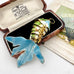 A decorative fish-shaped pendant  with a blue tail and green-and-white scaled body, displayed in an open vintage jewelry box. The box lid shows printed branding text, and part of a fan illustration is visible on the right.