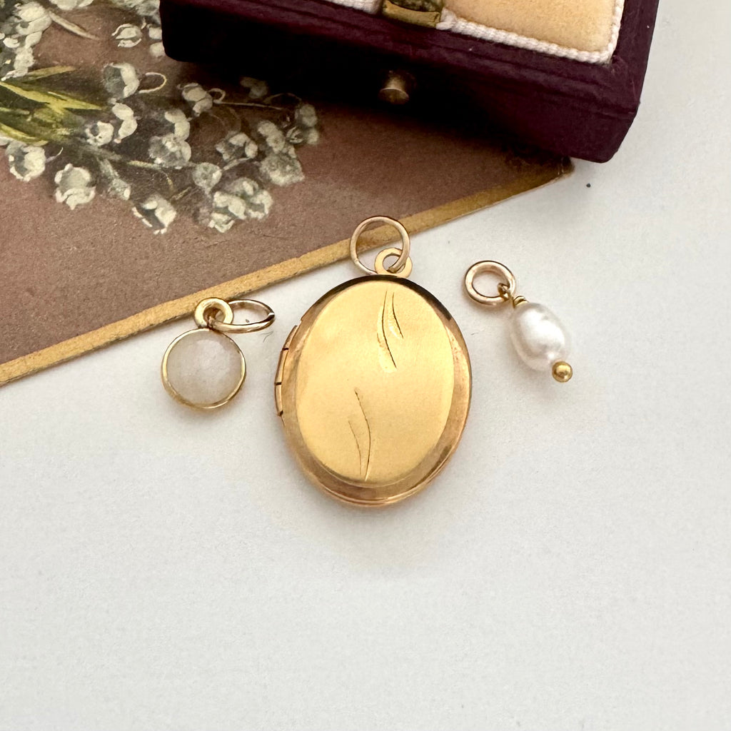Gold locket with leaf design and a pearl and moonstone charm on a white surface.