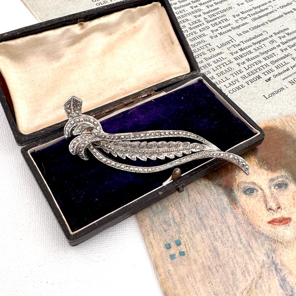 Silver brooch with decorative design in an open jewelry box on a textured surface.