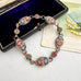 An ornate vintage micro mosaic bracelet with large oval and round enamel medallions in red, blue, white, and gold, displayed on a white textured surface next to an open dark velvet jewelry box and a decorative card.