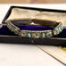 A vintage micro mosaic bracelet with green and white floral enamel inlays, displayed inside an open vintage presentation box with a purple velvet lining.