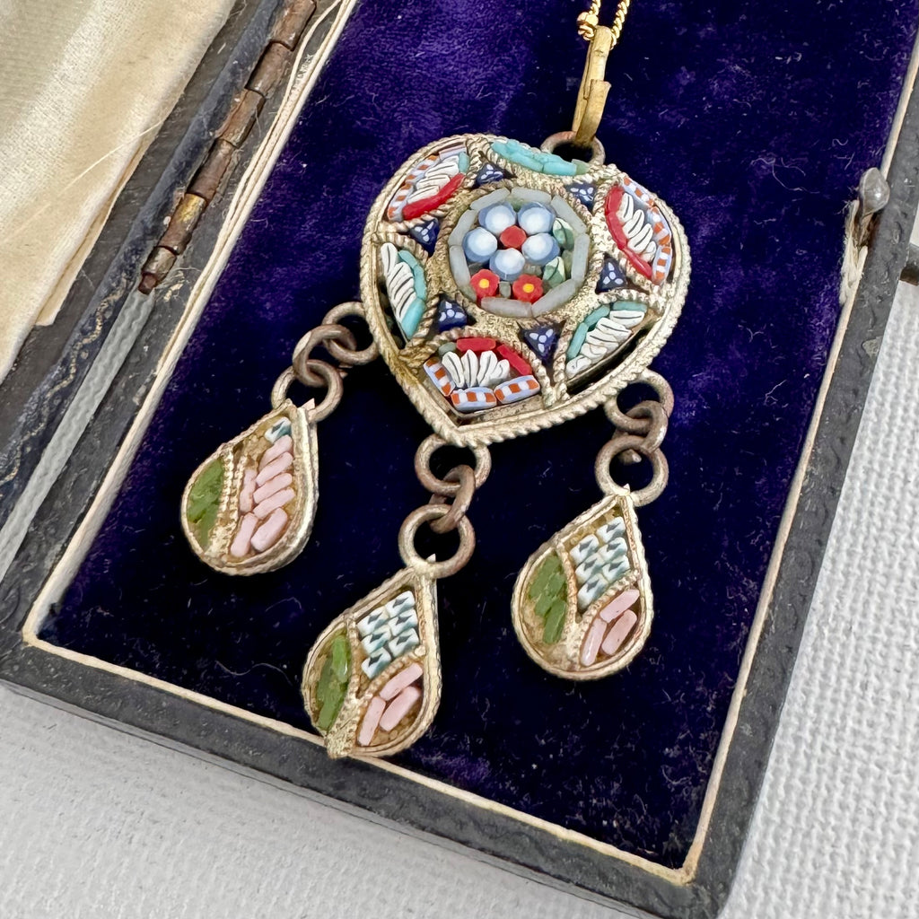 An ornate gold-toned pendant necklace with three dangling teardrop charms, displayed inside an open dark box lined with deep purple velvet. The pendant features intricate multicolor enamel-style mosaic details in red, turquoise, green, white, and pink.