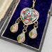 An ornate gold-toned pendant necklace with three dangling teardrop charms, displayed inside an open dark box lined with deep purple velvet. The pendant features intricate multicolor enamel-style mosaic details in red, turquoise, green, white, and pink.