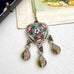 An ornate vintage-style pendant necklace with a gold-tone frame and three dangling teardrop charms, decorated with colorful mosaic and enamel-like patterns in pink, blue, green, red, and white, displayed on a textured white surface.