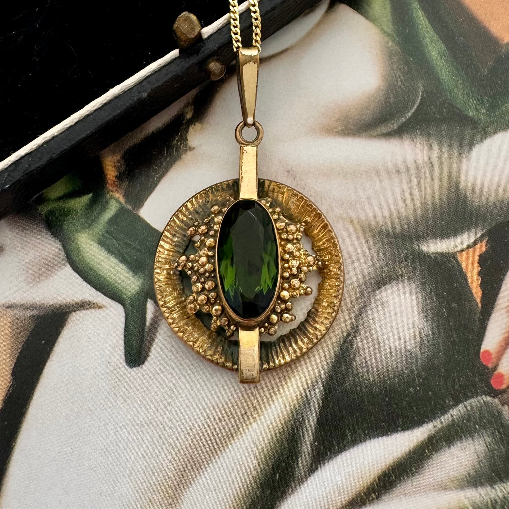 A vintage Andrea Daub gold pendant necklace featuring a large oval dark green gemstone surrounded by textured gold beads and a circular gold frame, resting on a vintage illustrated card.
