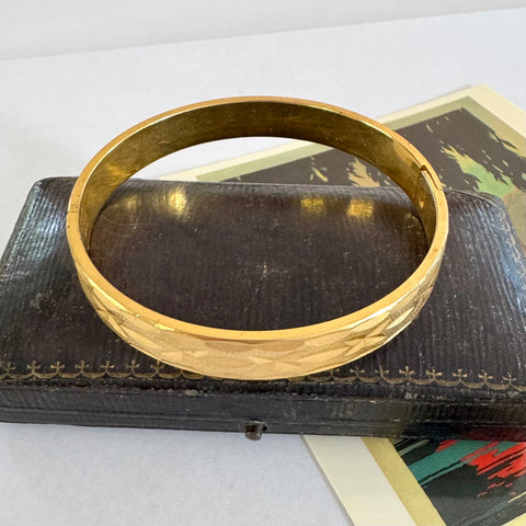 A polished gold bangle bracelet with a textured, diamond-patterned outer band, displayed on top of a dark vintage leather case.