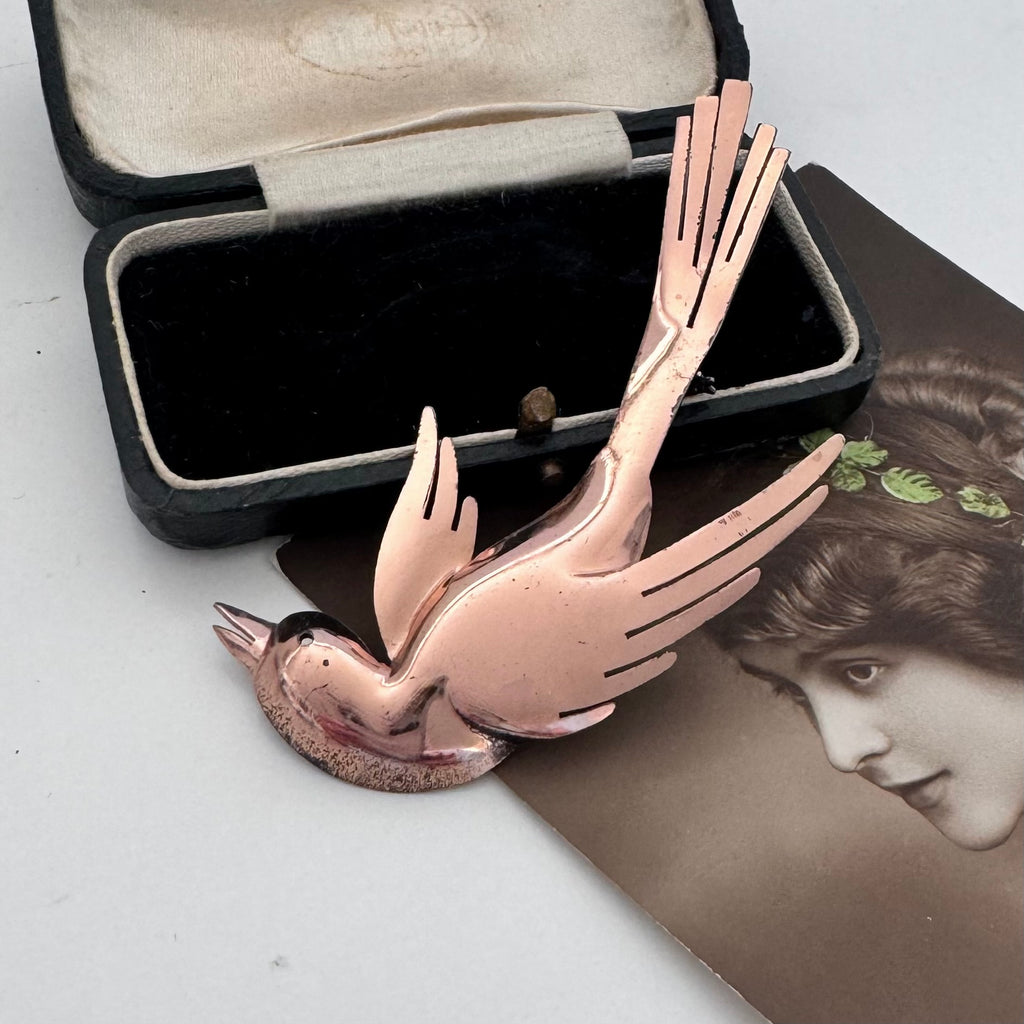 A vintage rose gold bird-shaped brooch with a polished metallic finish, displayed in front of its black velvet jewelry case and a vintage sepia-toned portrait card.