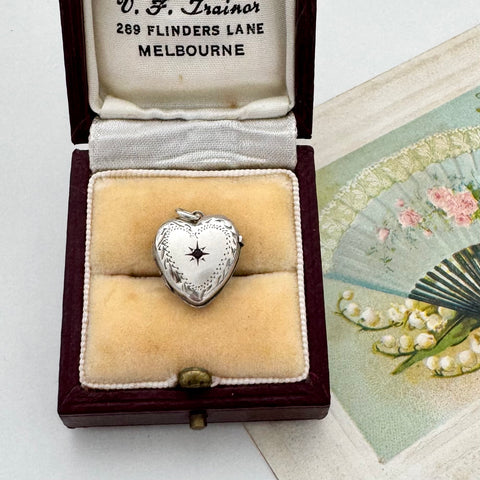 Silver heart-shaped locket in an open jewelry box with a fan and floral card in the background.