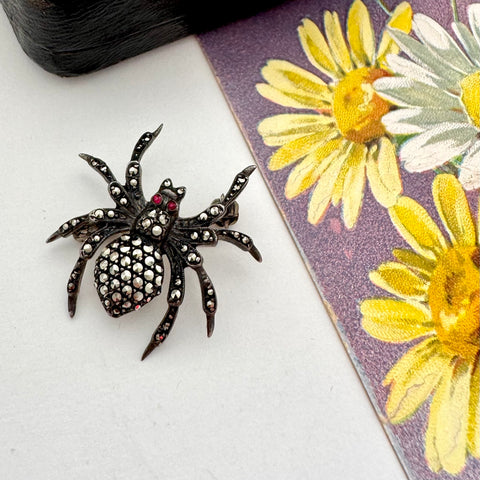 Black spider-shaped brooch on a white surface with a floral book in the background