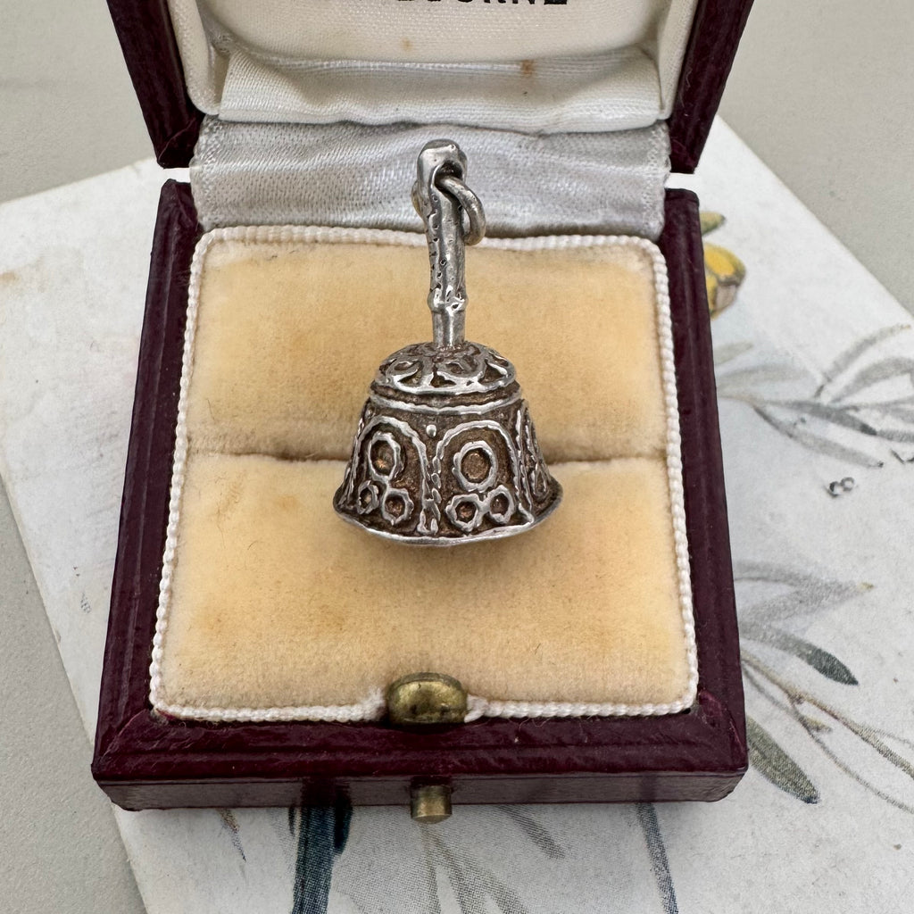 A vintage silver bell-shaped charm with intricate filigree cutouts and a hook at the top, displayed inside an open burgundy jewelry box with a cream velvet lining.
