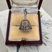 A vintage silver bell-shaped charm with intricate filigree cutouts and a hook at the top, displayed inside an open burgundy jewelry box with a cream velvet lining.