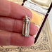 Silver column-shaped charm held between fingers with an ornate box in the background