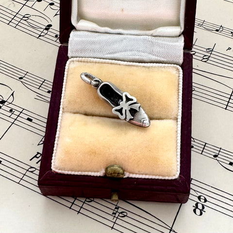 Silver shoe charm with a bow on a beige pillow in an open jewelry box on sheet music.